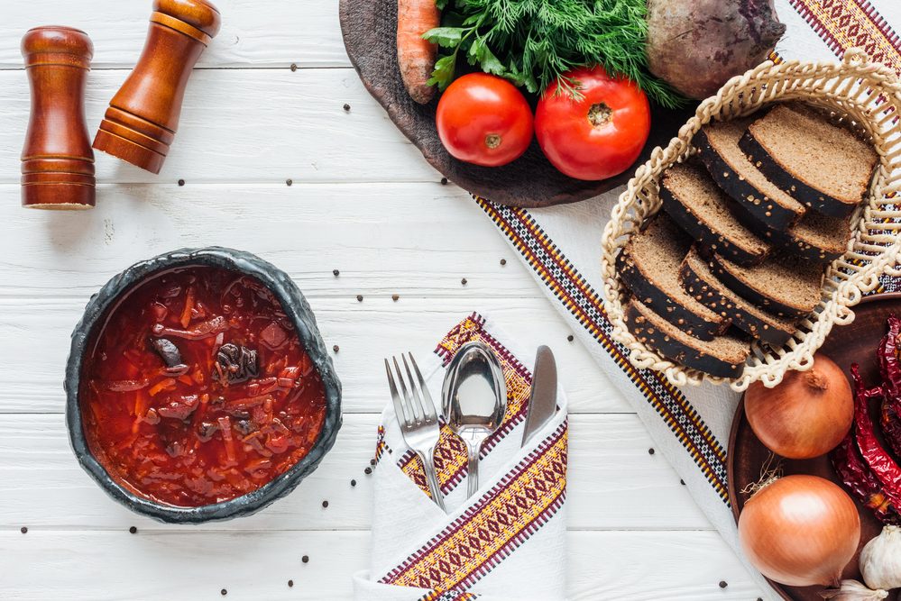 Борщ в глиняной миске с чёрным хлебом, овощами и вышитым рушником на белом столе